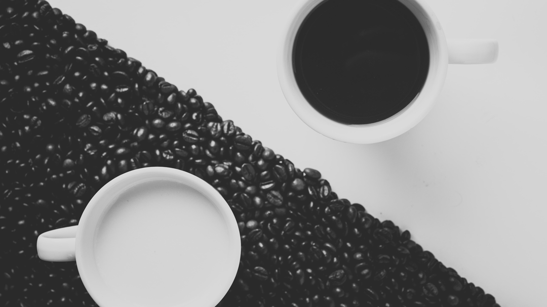 Coffee cups with one filled of white on top of black beans and the other filled with black in top of white surface