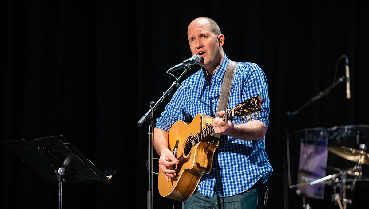 Man in blue button up shirt playing guitar and singing alongside the worship team on stage